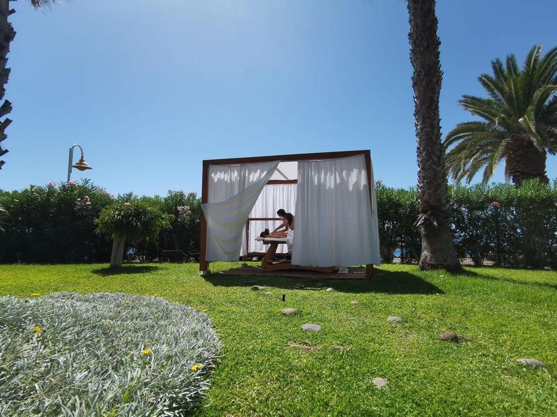Jardín con césped verde, pabellón con cortinas y persona debajo, palmeras al fondo.