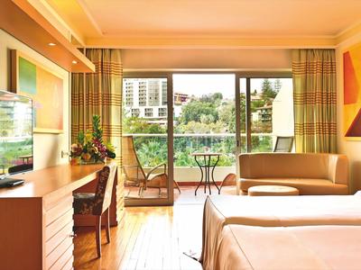 Hell eingerichtetes Hotelzimmer mit Doppelbett, Balkon und Blick auf eine Stadtlandschaft.