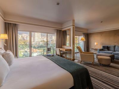 Modernes Hotelzimmer mit großem Bett, Sitzbereich und Panoramafenstern mit Stadtblick.