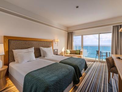 Helles Hotelzimmer mit zwei Betten, Sitzbereich und Blick aufs Meer durch große Fenster.