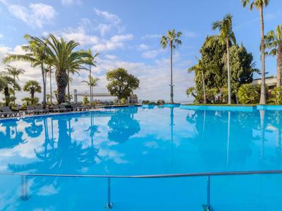 Großer blauer Pool mit Palmen und Liegestühlen unter bewölktem Himmel.