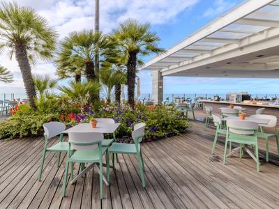 Hotelterrasse mit Holzboden, grünen Stühlen und Tischen, Blick auf Palmen und Meer.