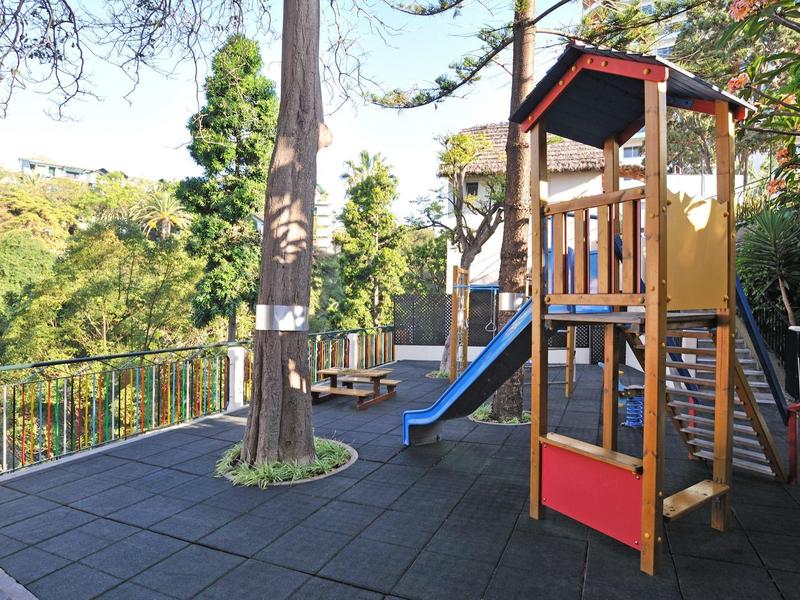 Spielplatz mit Klettergerüst, blauer Rutsche und Baum auf schwarzem Gummiboden im Freien.