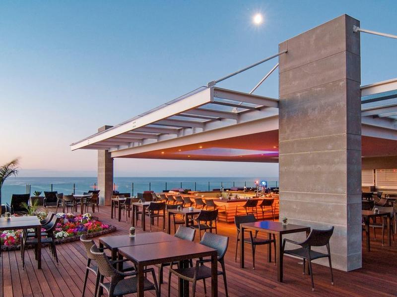 Offenes Restaurant mit Holzboden, Stühlen und Tischen unter Glasdach, Meerblick und Abendhimmel.