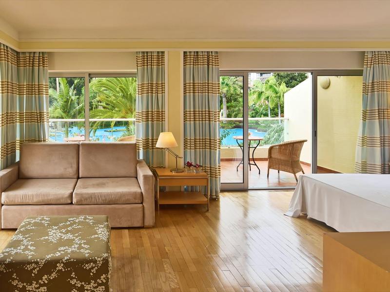 Helles Hotelzimmer mit Sofa, Bett, Nachttisch, Lampe und Zugang zu Terrasse mit Gartenblick.