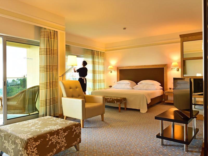 Helles Hotelzimmer mit Doppelbett, Stuhl, Fernseher, Balkon und warmem Licht.