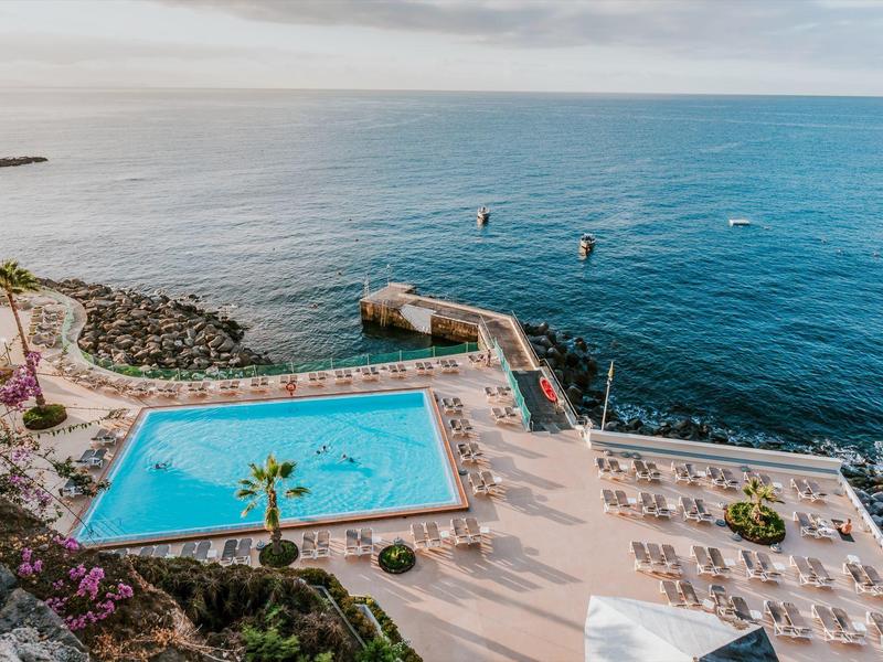 Blick auf ein blaues Poolbecken am Meer mit sonnigem Strandbereich und Steg ins Wasser.