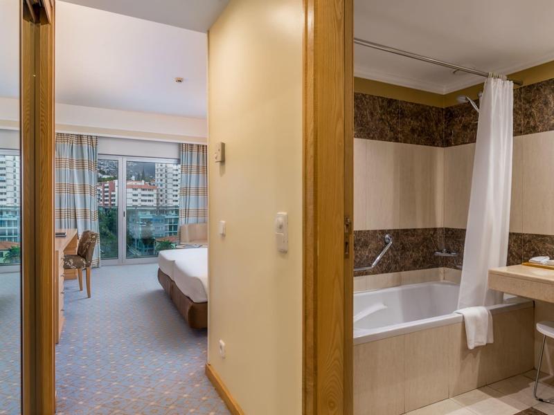 Ein helles Hotelzimmer mit Bett, Balkon und angrenzendem Badezimmer mit Badewanne.