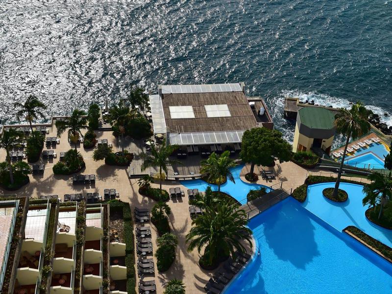 Hotelpool mit blauer Wasserfläche, Sonnenschirmen, Palmen und Blick auf das glänzende Meer.