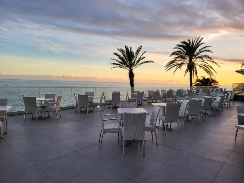 Leerer Außenbereich eines Restaurants mit weißen Tischen und Stühlen, Palmen und Meer im Hintergrund