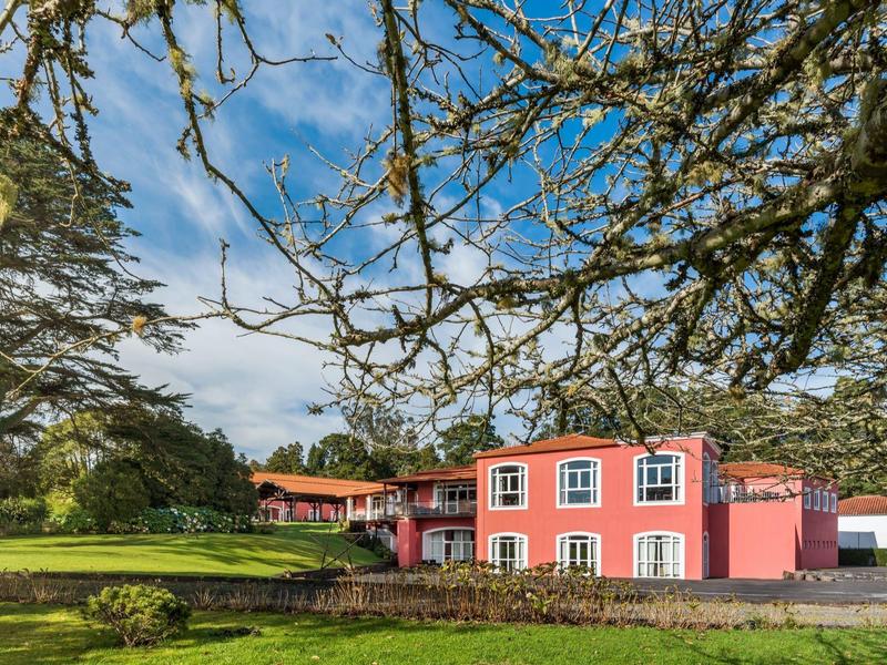 Růžový dům s bílými okny stojí na zeleném trávníku pod modrou oblohou.