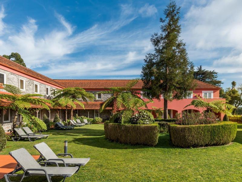 Hotelová zahrada se zelenými trávníky, lehátky a obklopena budovami s červenými střechami.