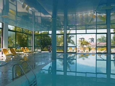 Innenpool mit großen Fensterfronten, blauer Wasseroberfläche, Liegestühlen und Außenblick.