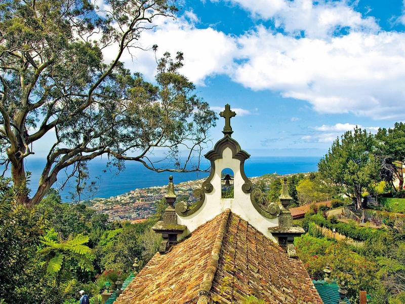 Dak van een gebouw met kruis in een tuin met uitzicht op zee onder een heldere hemel.