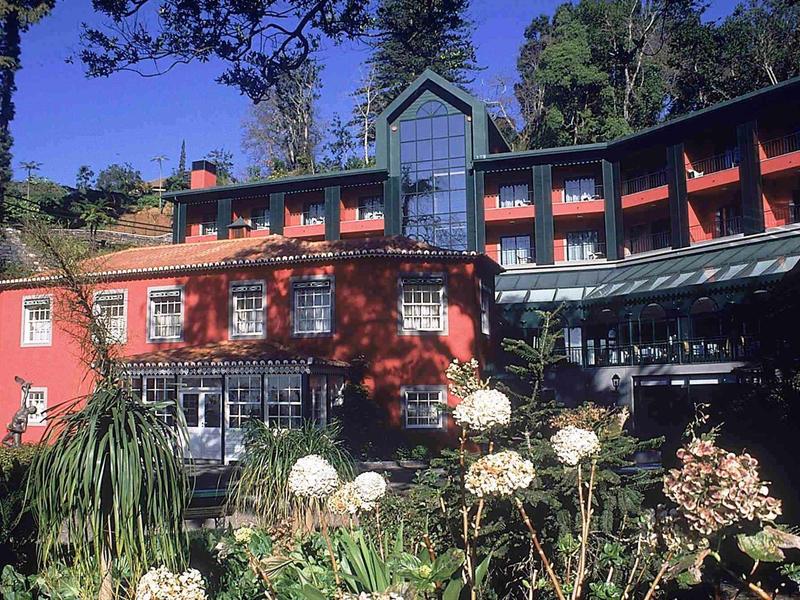 Een groot, gebogen rood hotel omgeven door bomen en bloementuin onder een blauwe lucht