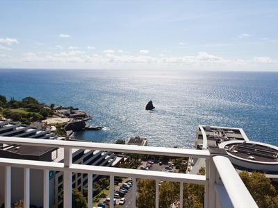 Blick von einem Balkon auf das blaue Meer, einen Felsen im Wasser und eine Küstenanlage bei Sonnenschein.