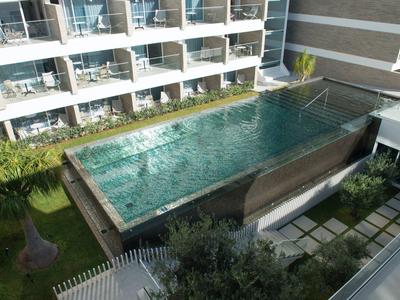 Moderner Glas-Pool auf Holzterrasse, umgeben von Grün, neben Wohngebäuden mit Balkonen.