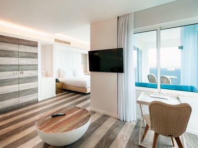 Modern eingerichteter Raum mit Holzboden, Bett, TV, Schrank und kleinem Tisch mit Stühlen am Fenster.