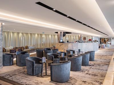 Großer, heller Hotel-Lobbybereich mit modernen grauen Sesseln und runden Tischen auf Teppichboden.