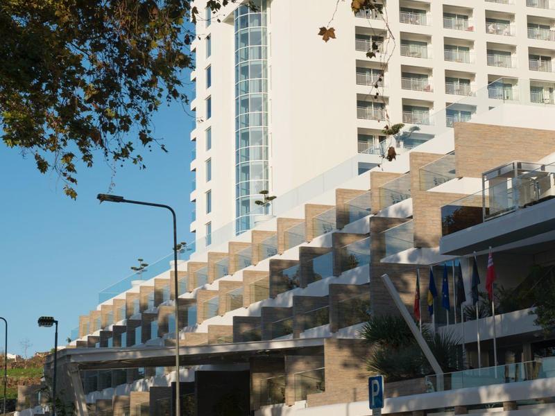 Velký moderní hotel s terasami, stromy a jasná modrá obloha.