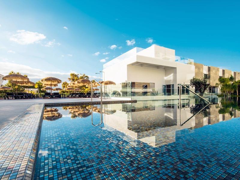 Klidný bazén s modrými dlaždicemi, odraz moderní bílé budovy a jasná obloha.