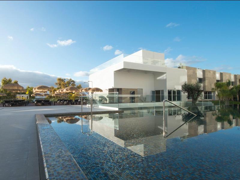 Moderní hotel s bazénem a odrazem pod modrou oblohou.