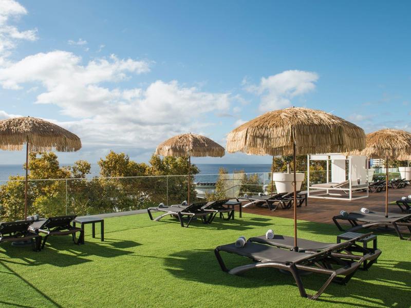 Slunečná terasa s lehátky pod slaměnými slunečníky s výhledem na oceán.