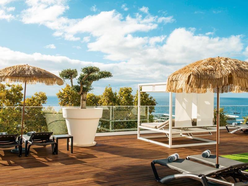 Dřevěná terasa s lehátky, slunečníky z trávy a výhledem na moře pod částečně zataženou oblohou.