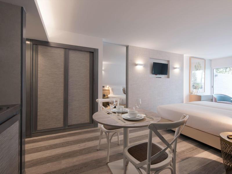 Světelný hotelový pokoj s manželskou postelí, kulatým stolem a velkými okny.