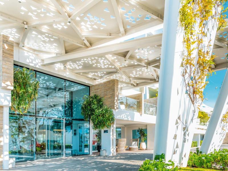 Moderní vstup do hotelu s skleněnou architekturou a stíny listí na bílých zdech.