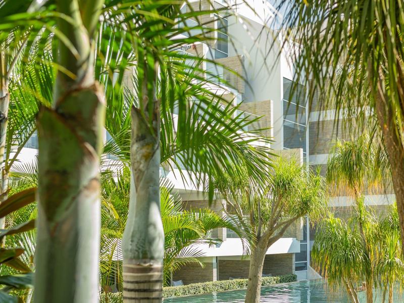 Bujné zelené palmy kolem bazénu s moderní budovou v pozadí.