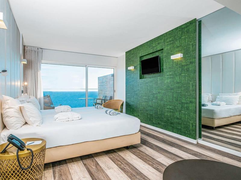 Moderní hotelový pokoj s manželskou postelí, výhledem na moře, zelenou zdí a dřevěnou podlahou.