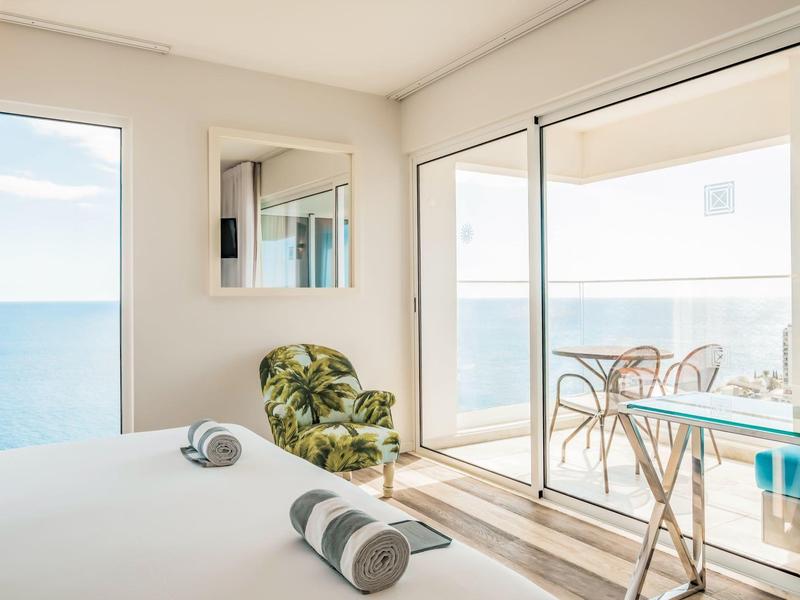 Světý hotelový pokoj s výhledem na moře, zelené křeslo a terasa s stolem a židlemi.