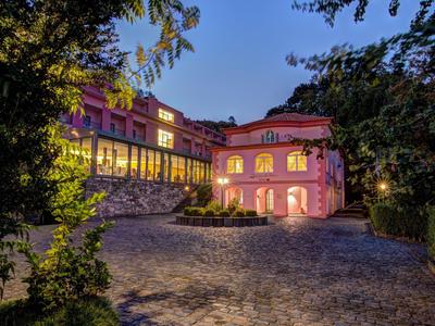 Großes Gebäude in Rosa mit vielen Fenstern, umgeben von Bäumen, beleuchtet in der Abenddämmerung.