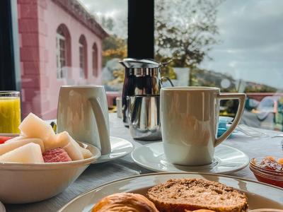 Frühstück mit Croissants, Marmelade, Obst, Kaffee und Saft an einem Tisch vor Fenster mit Blick.