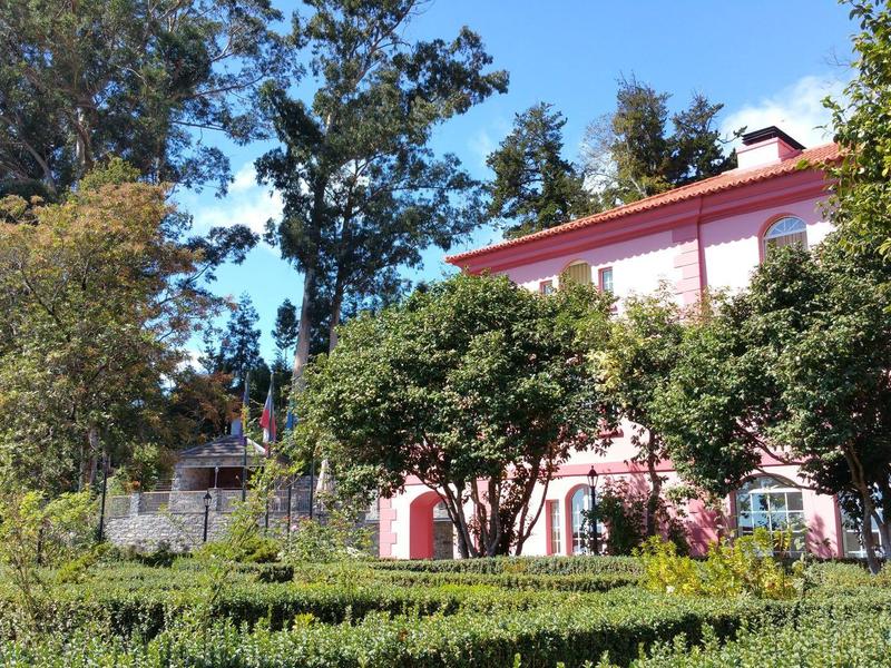 Gebäude mit rosa Fassade, umgeben von grünen Bäumen und gepflegtem Heckenlabyrinth im Sonnenschein.