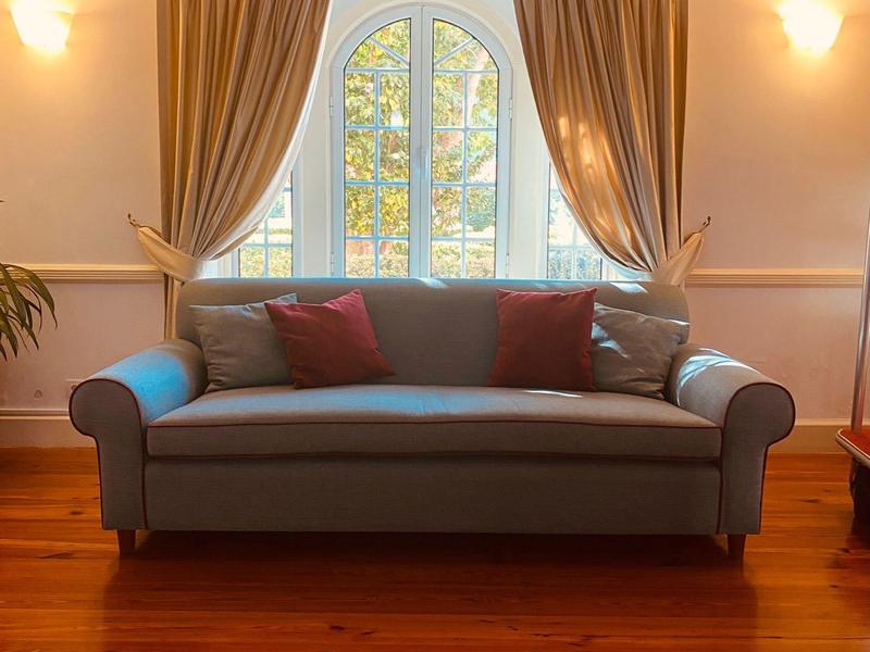 Ein blaugraues Sofa mit roten Kissen vor einem Rundfenster mit hellbraunen Vorhängen.