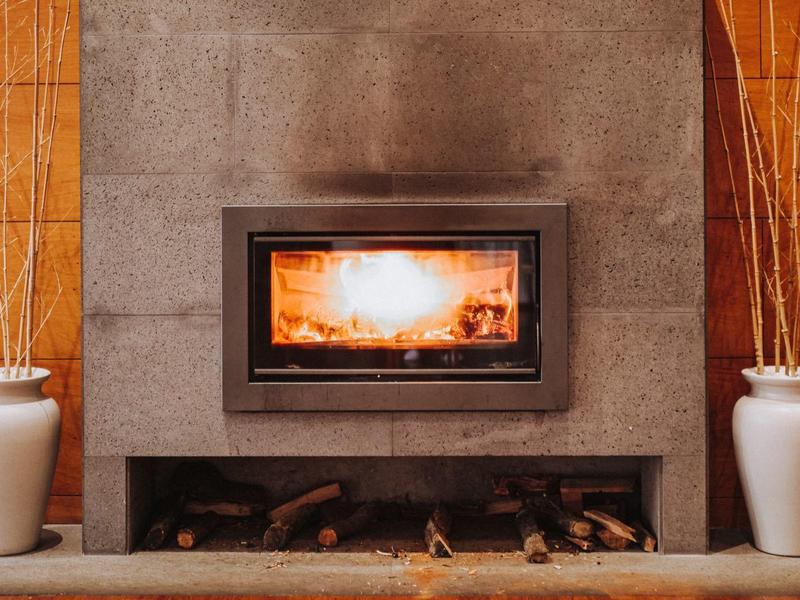 Moderner Kamin mit Glasfront in Wand integriert, davor Holzlager, links und rechts weiße Vasen mit Zweigen.