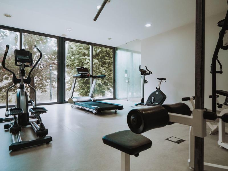 Helles Fitnessstudio mit großen Fenstern, Laufband, Heimtrainer und Kraftgerät auf hellem Boden.