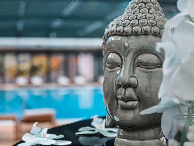 Graue Buddha-Statue vor weißem Orchideenstrauß, unscharf blauer Pool und Holzmöbel im Hintergrund.