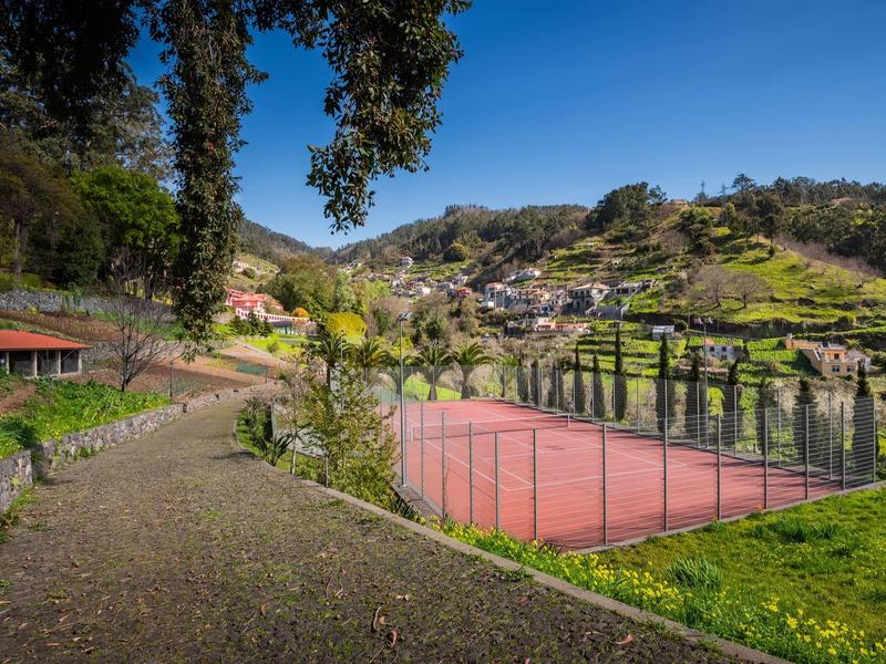 Roter Tennisplatz an einem Hügel, umgeben von grünem Gras und Bäumen, mit Häusern im Hintergrund.