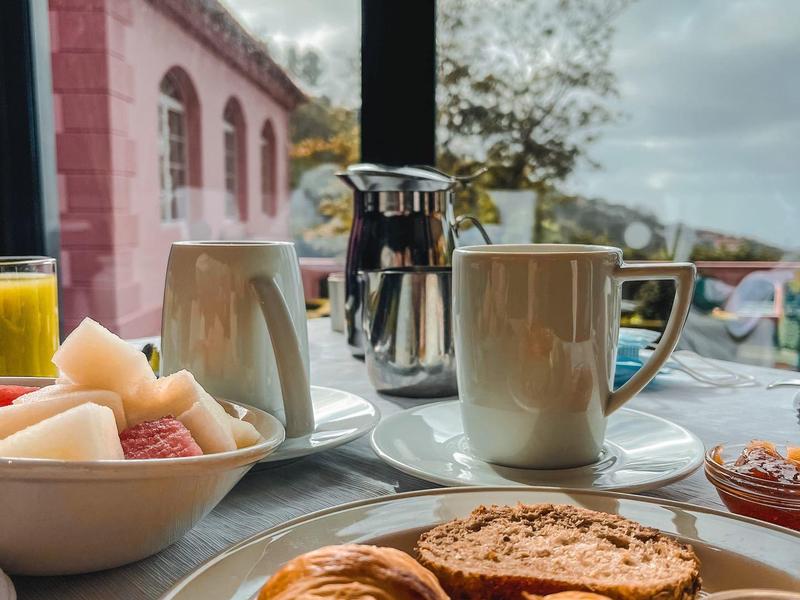 Frühstück mit Croissants, Marmelade, Obst, Kaffee und Saft an einem Tisch vor Fenster mit Blick.