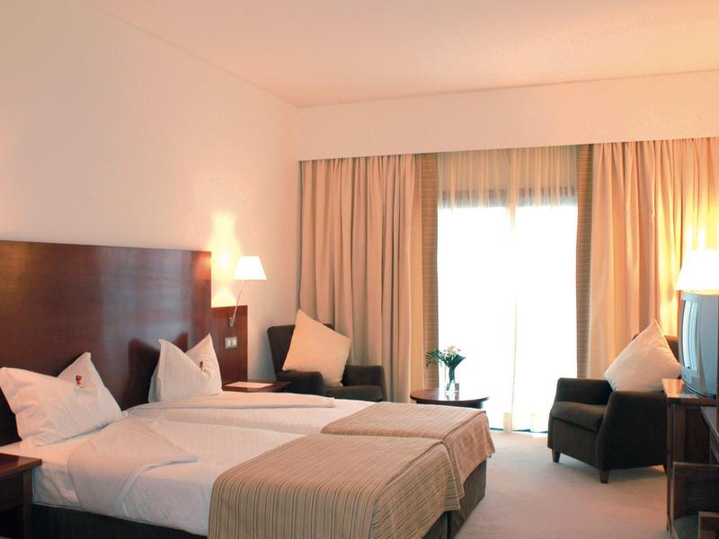 Helles Hotelzimmer mit zwei Betten, beigen Vorhängen, Sesseln und einem kleinen Tisch vor dem Fenster.