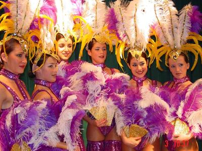 Sechs Frauen in bunten Federkostümen posieren vor einer beleuchteten Kulisse.