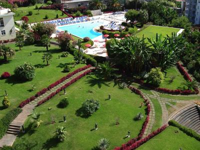 Zelený hotelový areál s bazénem a dlážděnými cestami obklopený květinami a keři.