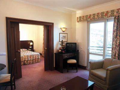 Světý hotelový pokoj s posezením, psacím stolem, televizí a výhledem do ložnice.