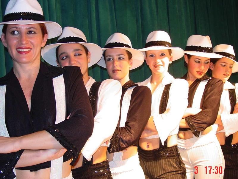 Šest žen v černo-bílém oblečení a kloboucích stojí před zeleným pozadím.