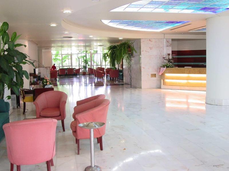 Světlá hotelová hala s růžovými křesly, zelenými rostlinami a osvětlenou recepcí.