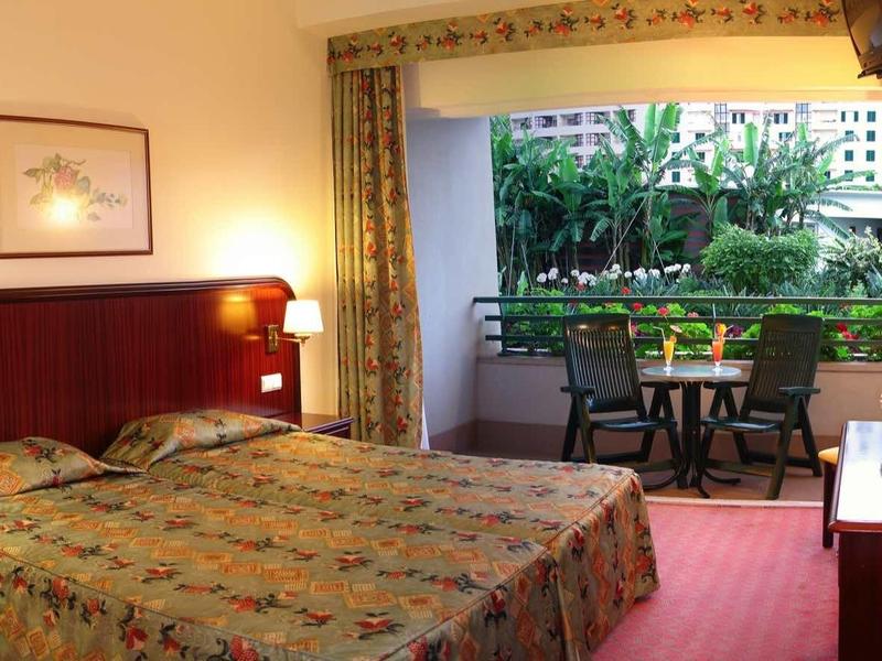 Pohodlný hotelový pokoj s manželskou postelí, psacím stolem a výhledem na zelenou terasu.