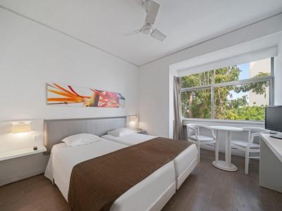 Moderní hotelový pokoj s manželskou postelí, velkým oknem, televizí a malým balkónem se židlemi.